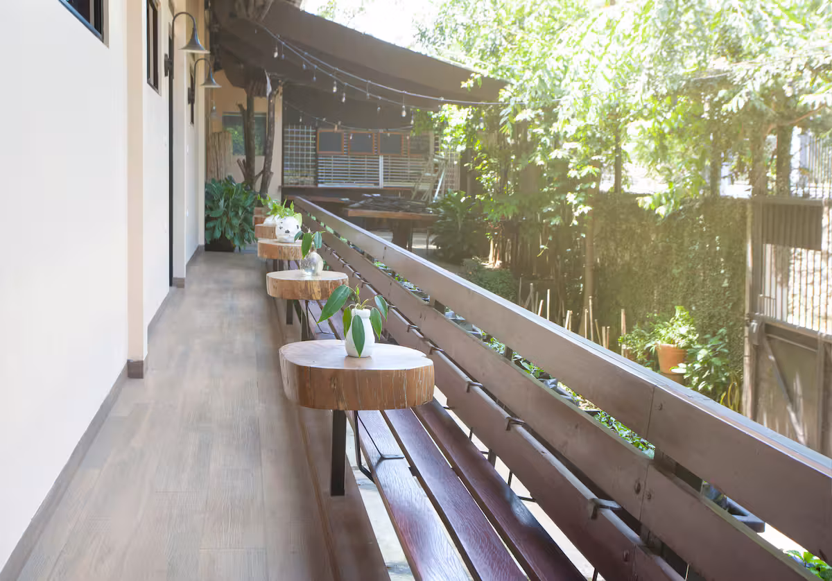 A calm wooden balcony with natural-wood tables, potted plants and string lights surrounded by greenery