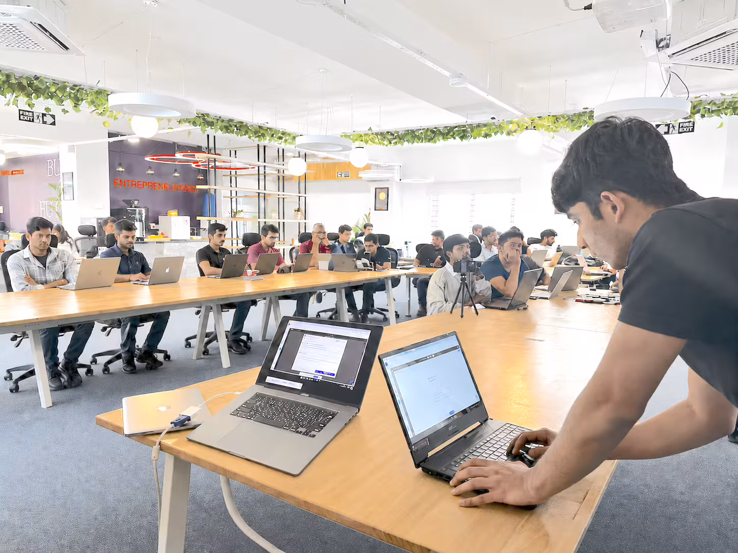 A room of learners working at laptops around wooden tables, greenery overhead